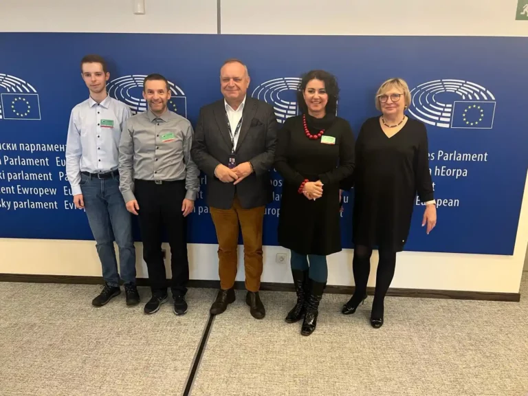 Grupa pięciu osób stoi przed niebieską ścianą z logo Parlamentu Europejskiego. Na ścianie widnieje napis "European Parliament" w kilku językach. Osoby są ubrane elegancko, w większości w ciemnych kolorach. Mężczyzna pośrodku ma na sobie ciemną marynarkę i brązowe spodnie, a kobieta obok niego nosi czarną sukienkę i czerwone korale. Pozostałe osoby również są elegancko ubrane. Wszyscy patrzą w stronę aparatu i uśmiechają się.
