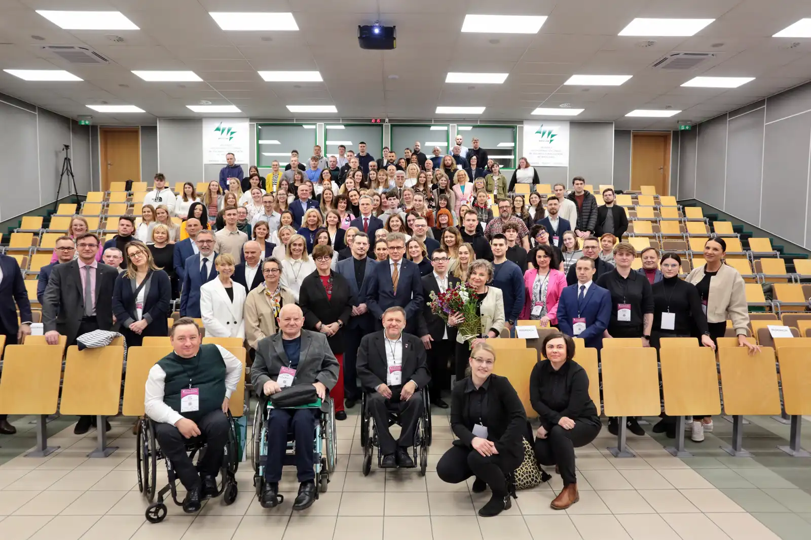 Zdjęcie przedstawia dużą grupę ludzi zgromadzonych w sali wykładowej. W tle widać rzędy pustych, drewnianych krzeseł. Na pierwszym planie znajduje się kilka osób na wózkach inwalidzkich oraz kobieta trzymająca bukiet kwiatów. Wszyscy uczestnicy są elegancko ubrani, co sugeruje, że może to być oficjalne wydarzenie lub konferencja. W tle, na ścianie, widoczne są dwa logotypy, które mogą być związane z organizacją wydarzenia. Sala jest dobrze oświetlona, a sufit pokryty jest panelami z oświetleniem.
