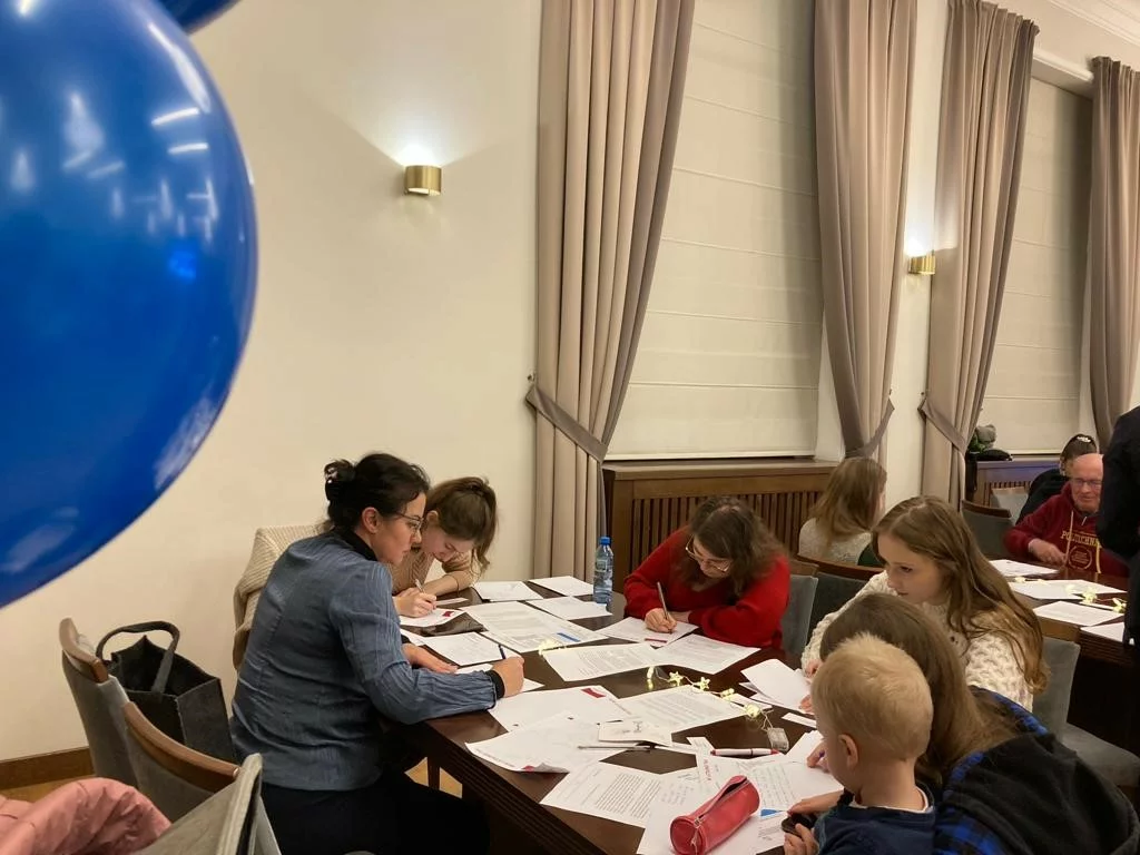 Grupa ludzi siedzących przy dużym stole w pomieszczeniu. Na stole leży wiele kartek papieru, a osoby są zajęte pisaniem. W tle widać zasłony i ścianę z lampami. Po lewej stronie zdjęcia widoczny jest duży niebieski balon. Wśród osób przy stole są zarówno dorośli, jak i dzieci. Atmosfera wydaje się być skupiona i spokojna.