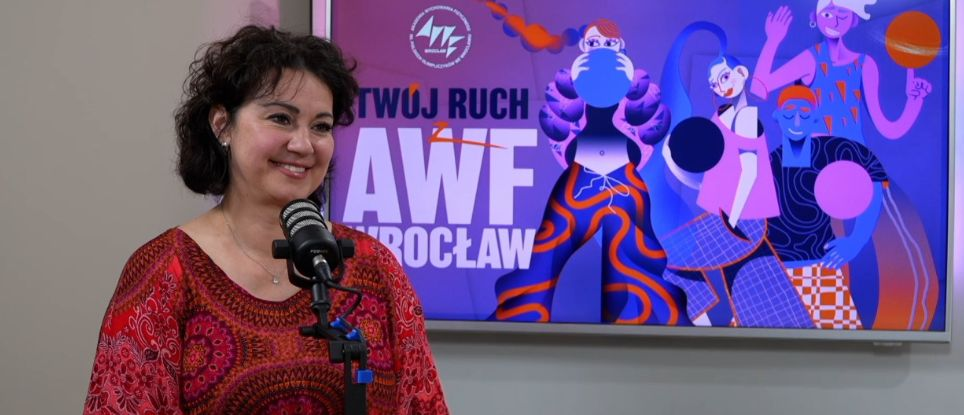 Dr Dominika Zawadzka siedzi przed mikrofonem. Ma na sobie czerwoną bluzkę z wzorami. W tle znajduje się kolorowy plakat z napisem "TWÓJ RUCH AWF WROCŁAW". Plakat przedstawia stylizowane postacie w dynamicznych pozach, trzymające różne sportowe akcesoria, takie jak piłki. Kolory plakatu są żywe, dominują odcienie fioletu, pomarańczu i niebieskiego.